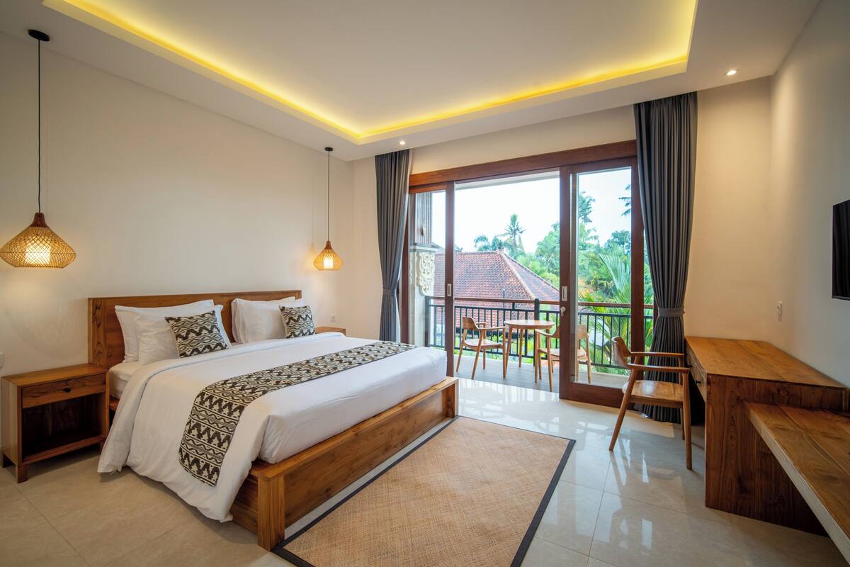 Een moderne slaapkamer met een groot bed, nachtkastjes, een bureau en een balkon met uitzicht op groen.