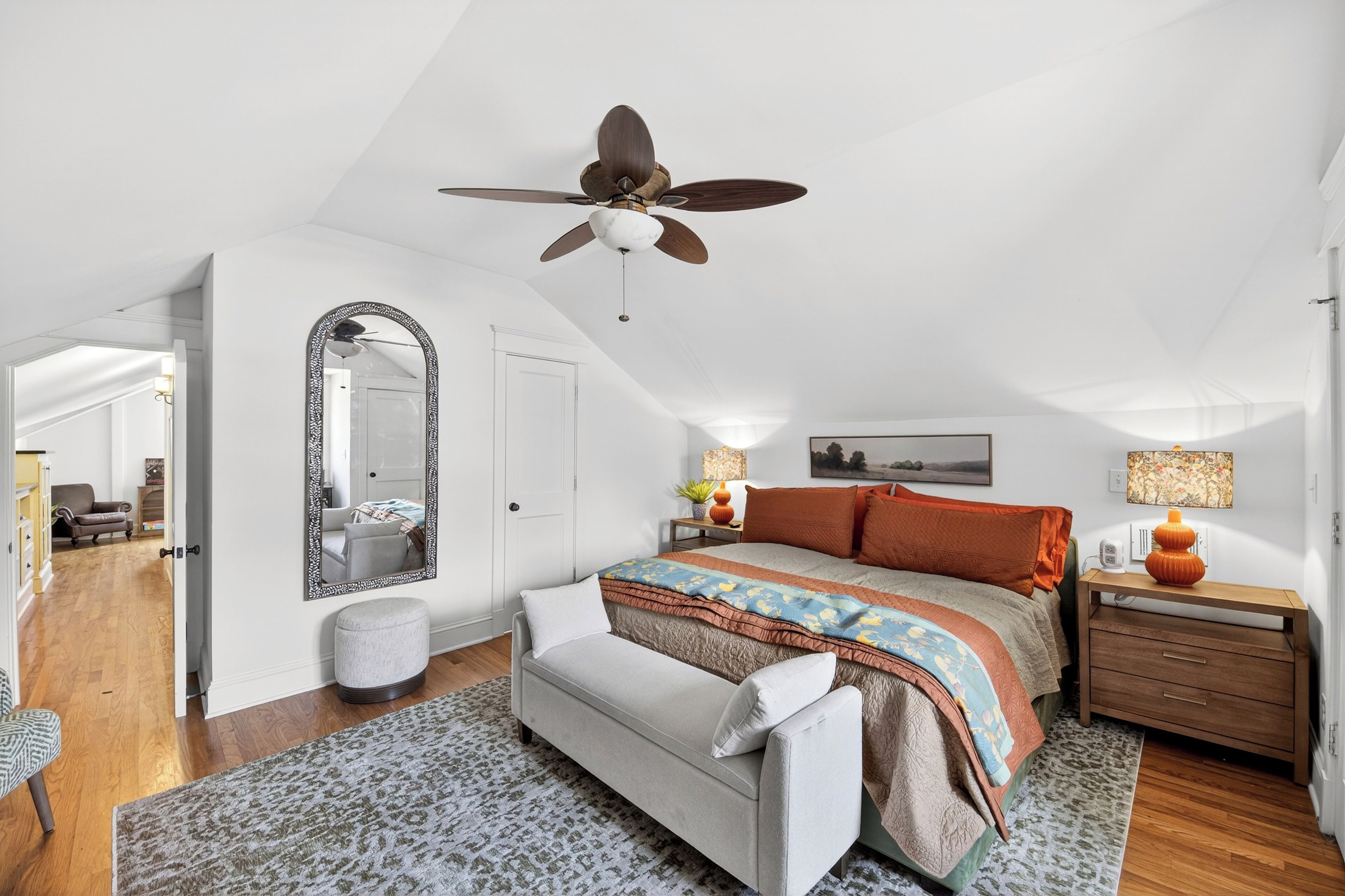 A bedroom with a large bed, a ceiling fan, a mirror, a nightstand, and a door leading to another room.