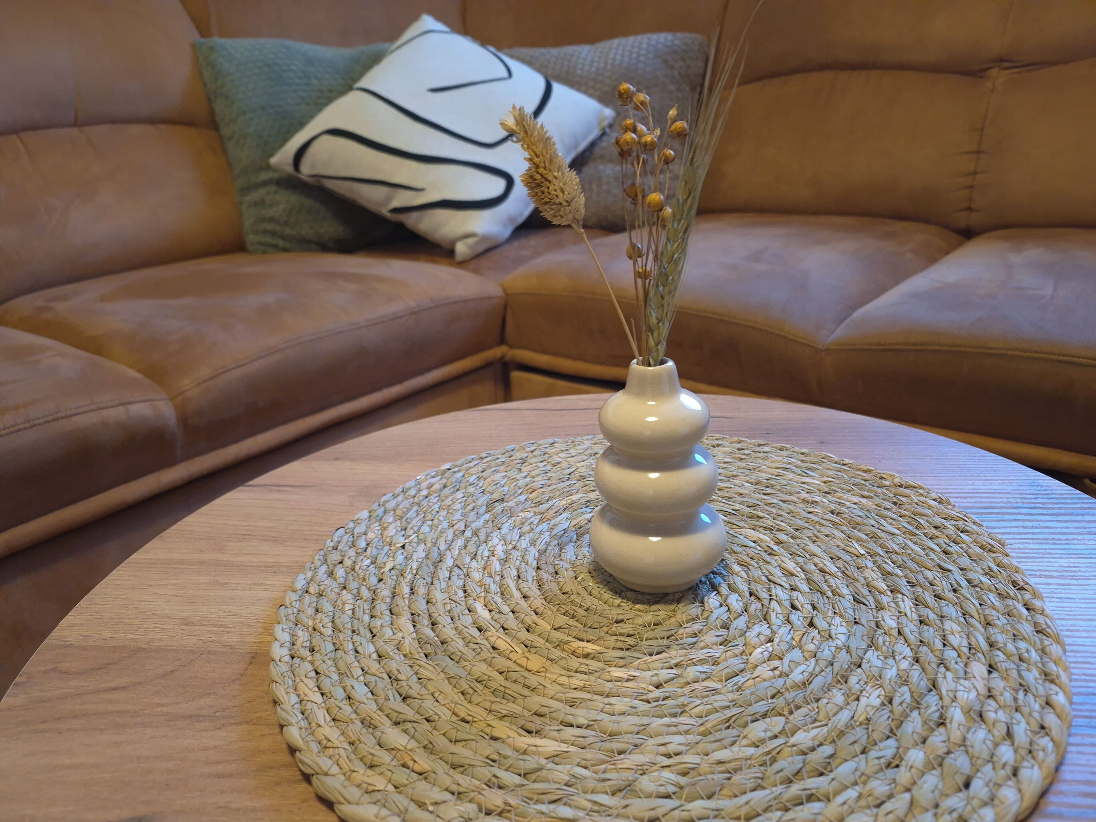 Ein runder Holztisch mit einer gewebten Tischunterlage, eine weiße Vase mit getrockneten Pflanzen und eine braune Ledersofa mit einem gemusterten Kissen im Hintergrund.