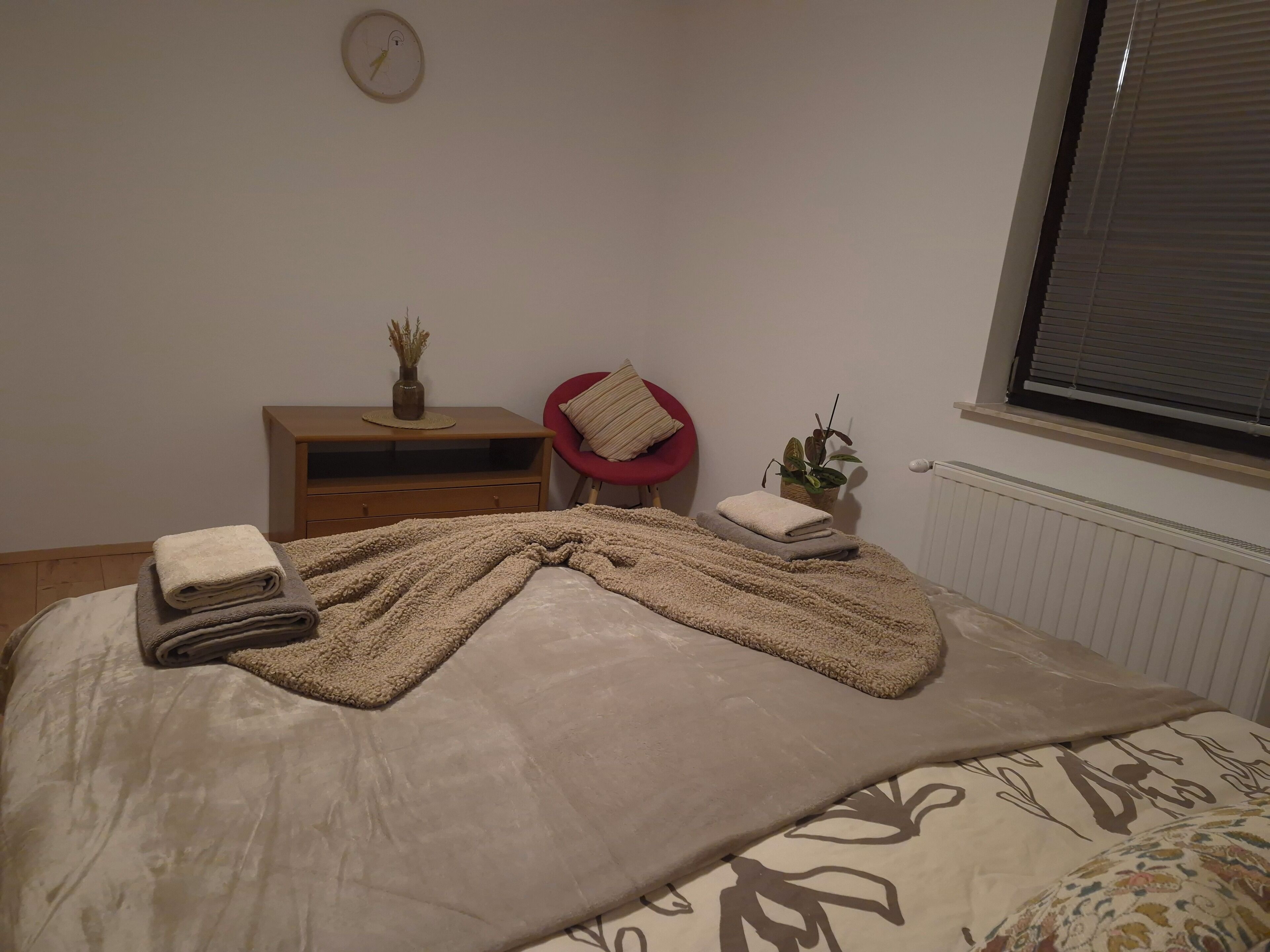 Ein ordentlich bezogenes Bett mit Decke und Handtüchern, ein kleiner Nachttisch mit Vase und Pflanze, ein roter Sessel und ein Fenster mit Jalousien.