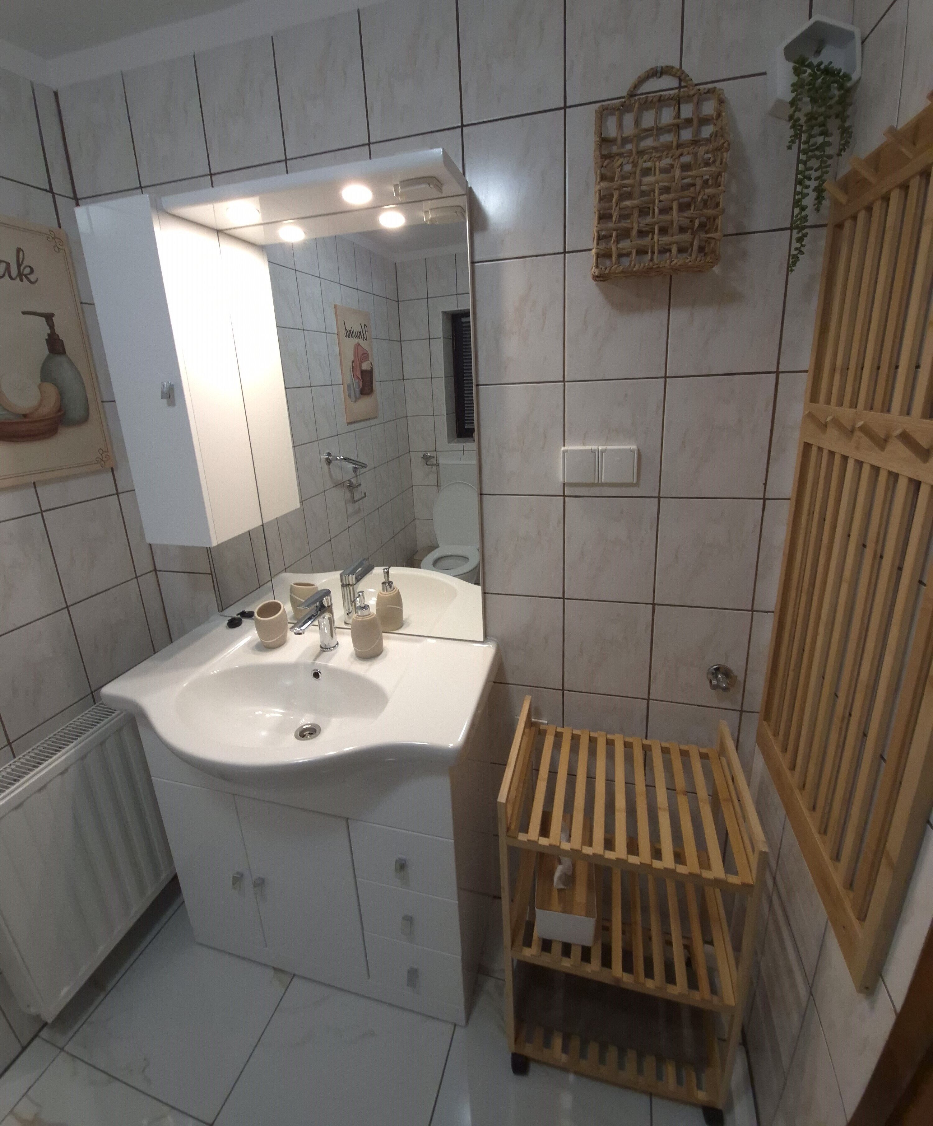 Ein Badezimmer mit Waschbecken, Spiegel und gefliesten Wänden. Ein Holzregal und eine hängende Pflanze sind ebenfalls vorhanden.
