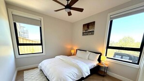 Un dormitorio con cama, dos lámparas de noche, ventilador de techo y ventanas amplias.
