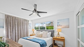 Ein modernes Schlafzimmer mit Bett, Nachttischen, Deckenventilator und Fenster mit Blick ins Grüne.