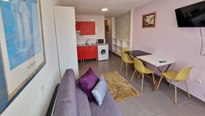 Un salon moderne comprenant un canapé, une table à manger avec des chaises, un coin cuisine avec des meubles rouges, un lave-linge et un téléviseur fixé au mur.