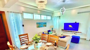 Ein modernes Wohnzimmer mit einem Glastisch, Holzsesseln, einer Couch, einem Fernseher und einem Bild an der Wand.