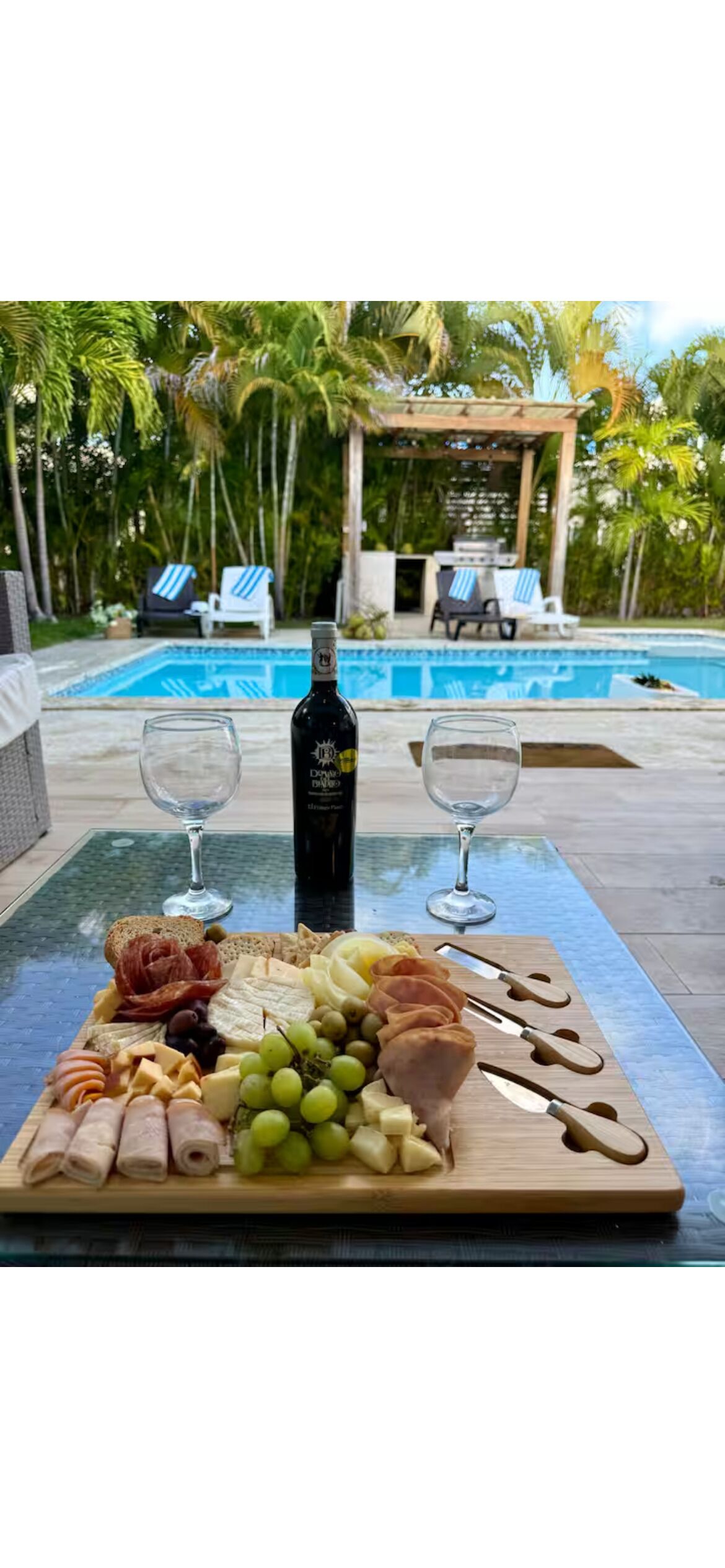 Una mesa con una tabla de embutidos, dos copas de vino y una botella, frente a una vista de piscina con sillones de descanso y palmeras.