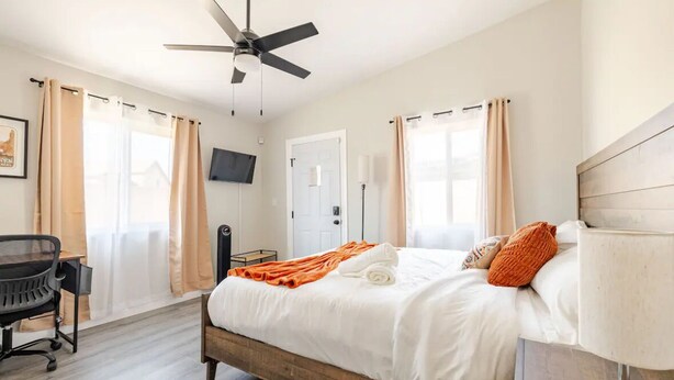 Une chambre à coucher avec un lit, un bureau, une chaise, un ventilateur de plafond et un téléviseur fixé au mur.