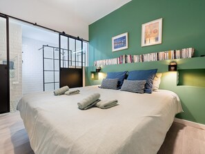 Een slaapkamer met een groot bed, een boekenplank vol boeken, twee ingelijste foto's en een schuifdeur naar een andere kamer.