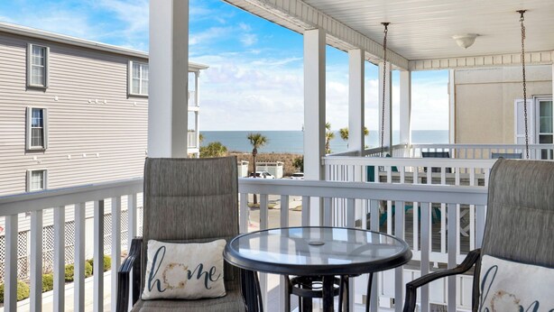 A covered balcony with two chairs and a table, overlooking a beach and ocean. - Easy Coastal Living Meets Relaxed Island Comfort | Heated Pool Access (Tybee Island)