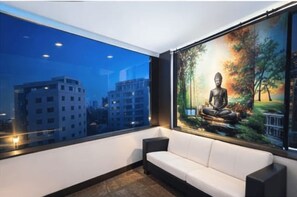 A modern interior with a white sofa, a large window with a cityscape view, and a Buddha painting. - At Homes Hotel Darbar (Hyderabad)