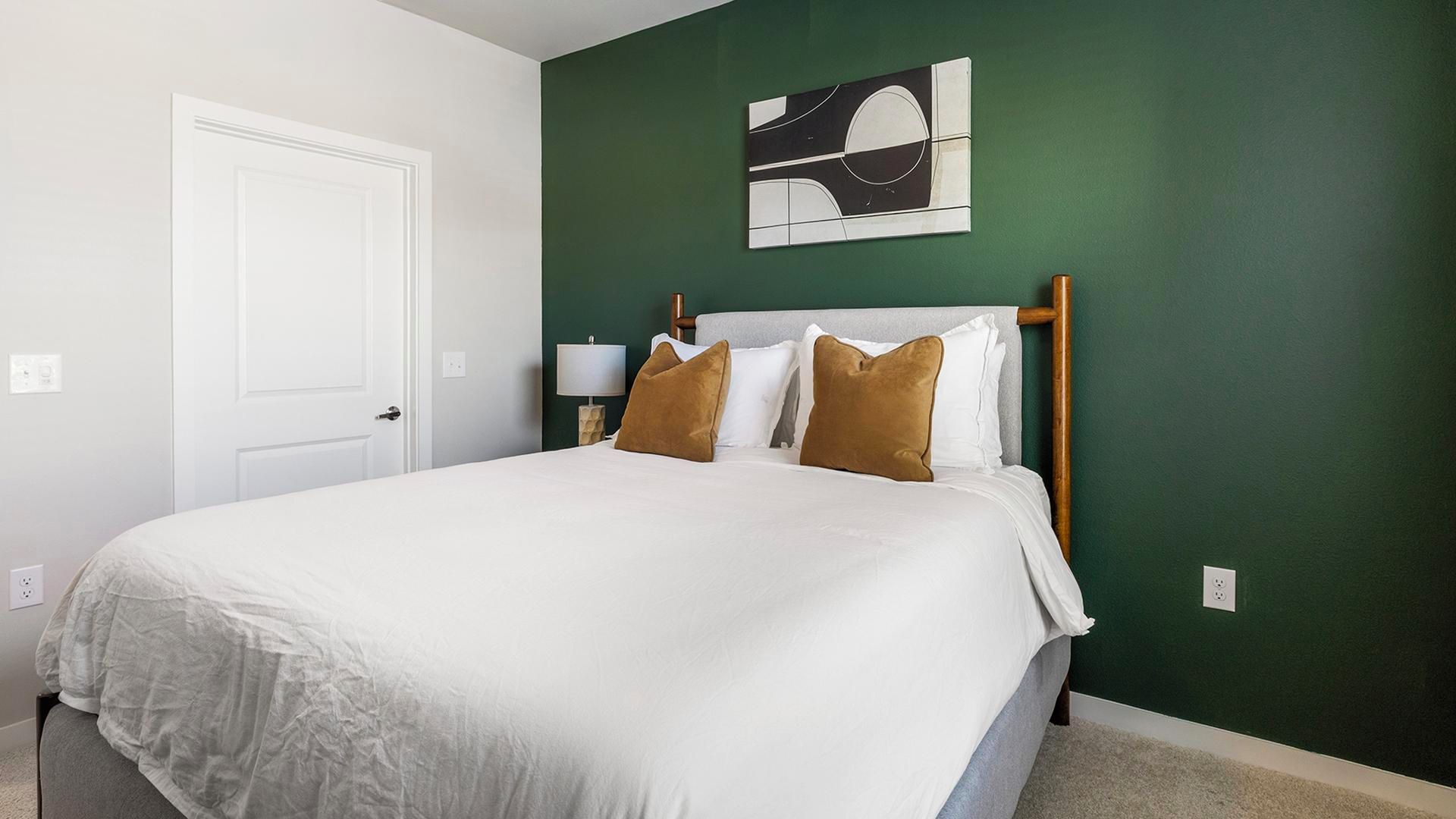 Een slaapkamer met een bed, kussens, een nachtkastje, een lamp en een schilderij aan de muur.