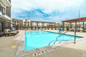 A swimming pool with lounge chairs, a pergola, and a building with multiple floors in the background. - Landing Furnished Apartments - Corinth (Denton)