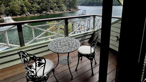 A covered outdoor deck with a table and chairs, overlooking a lake with boats and a forested hillside. - A Little Piece of Paradise – Cottage Point Waterfront Retreat (Cottage Point)