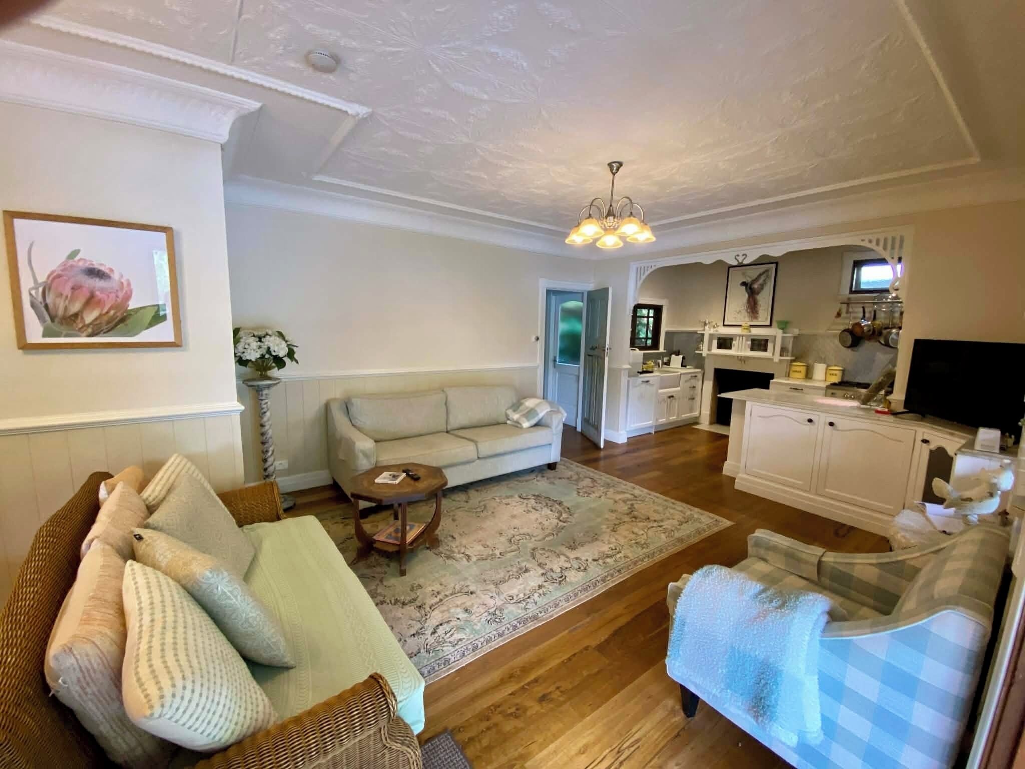 A living room with a sofa, a wicker chair, a small table, and a blue checkered chair. There is a kitchen in the background with cabinets, a stove, and a television.