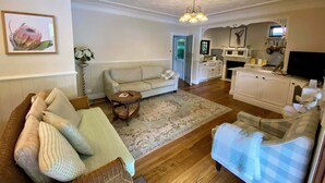 A living room with a sofa, a wicker chair, a small table, and a blue checkered chair. There is a kitchen in the background with cabinets, a stove, and a television. - A Little Piece of Paradise – Cottage Point Waterfront Retreat (Cottage Point)