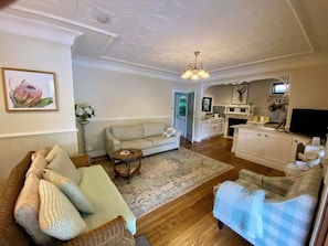 A living room with a sofa, a wicker chair, a small table, and a blue checkered chair. There is a kitchen in the background with cabinets, a stove, and a television. - A Little Piece of Paradise – Cottage Point Waterfront Retreat (Cottage Point)