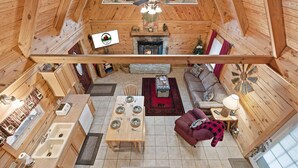Interior de uma cabana de madeira com área de jantar, lareira, sofá e ventilador de teto.