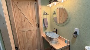 A bathroom with a wooden door, a round mirror, a sink with a metal basin, and a towel hanging on the wall. - The Cabin in Tribune (Tribune)
