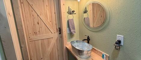 A bathroom with a wooden door, a round mirror, a sink with a metal basin, and a towel hanging on the wall.