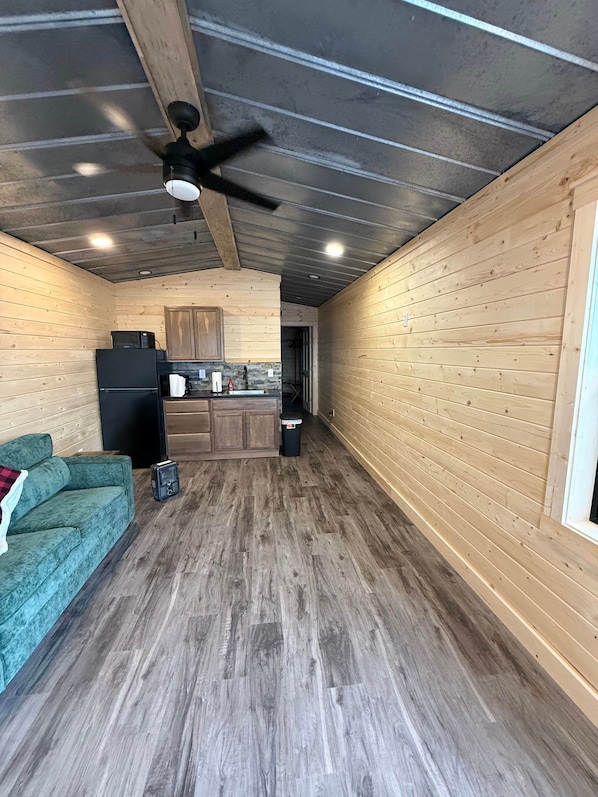 Un interior acogedor de cabaña con piso de madera, un sofá verde, una zona de cocina con armarios de madera y un ventilador de techo.