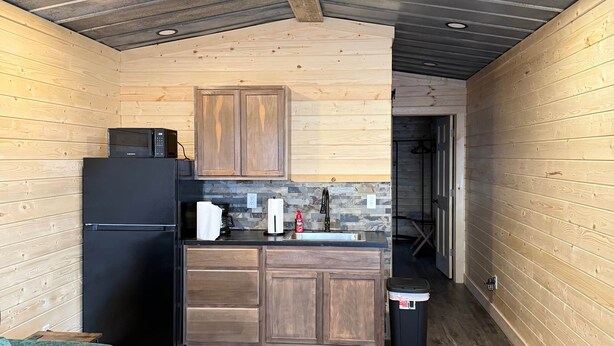 A compact kitchen with wooden cabinets, a black refrigerator, a microwave, and a sink. There's a ceiling fan, a small trash bin, and a teal sofa. - The Cabin in Tribune (Tribune)
