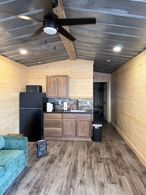 Una cocina compacta con armarios de madera, un refrigerador negro, un microondas y un lavabo. Hay un ventilador de techo, un cesto de basura pequeño y un sofá color turquesa.