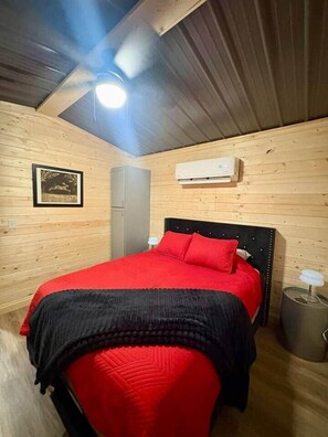 A cozy bedroom with a wooden ceiling, a bed with red bedding, a grey wardrobe, and a white air conditioning unit.