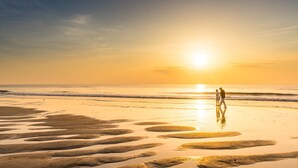 Une scène de plage au coucher du soleil, avec deux personnes qui se promènent et des motifs sur le sable mouillé.