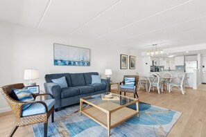 Ein modernes Wohnzimmer mit einer Couch, zwei Sesseln, einem Glastisch und einem Essbereich mit weißem Tisch und Stühlen.