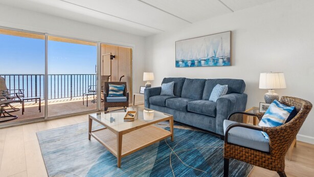Un salon avec un canapé, un fauteuil et une table basse, un balcon avec vue sur la mer, et un tableau accroché au mur.