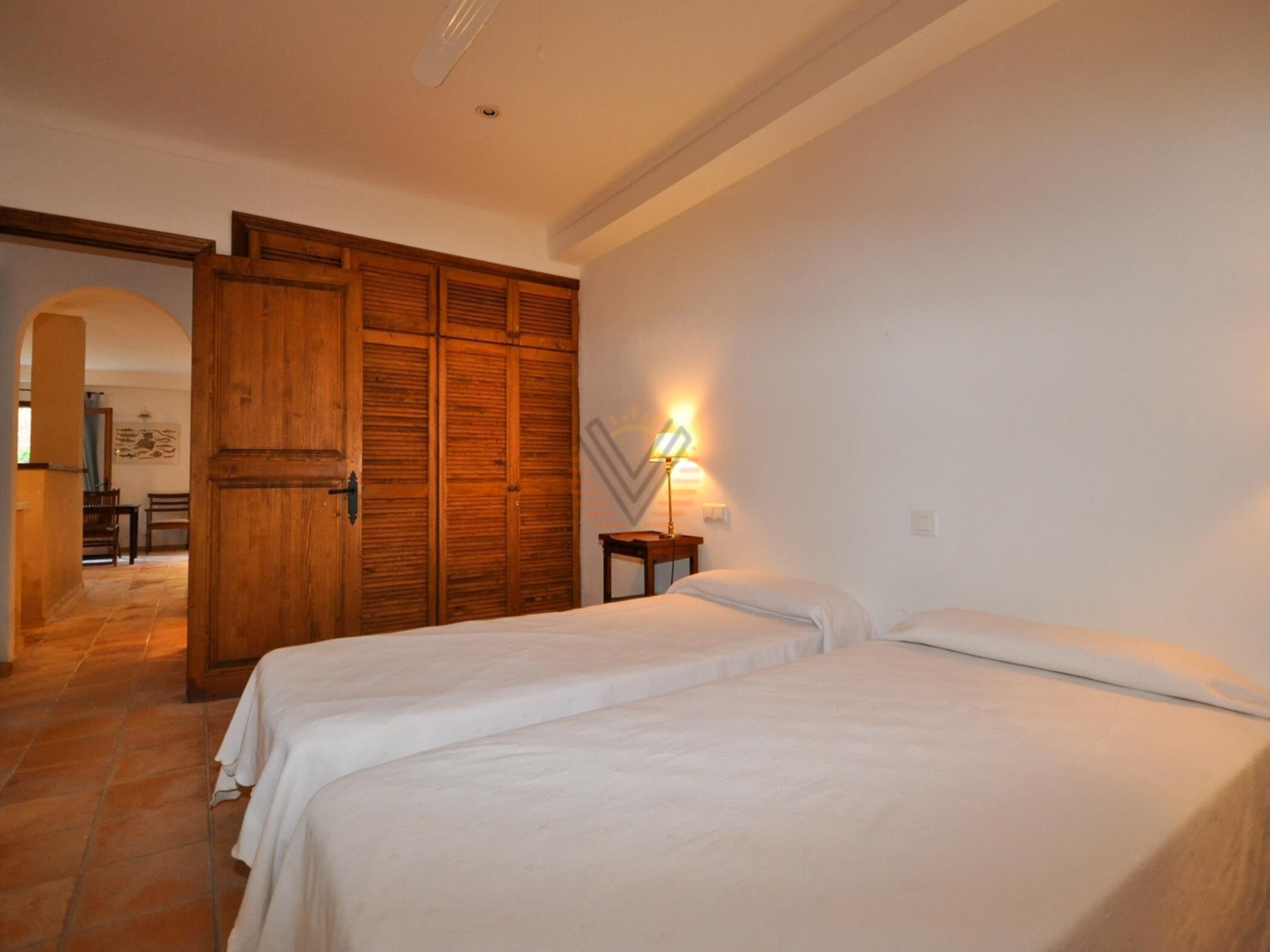 Una habitación con dos camas, un armario de madera, una mesita con lámpara y un acceso a otra habitación.
