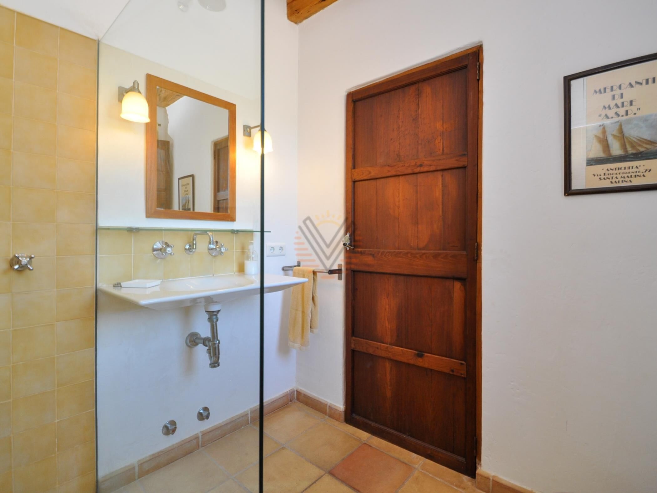 Um banheiro com porta de madeira, pia branca e espelho acima, e piso revestido de azulejos.
