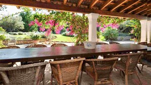 An outdoor covered patio with a long table and wicker chairs, surrounded by lush greenery and flowering plants. - Can Teulada, private pool Pollensa 8 persons (Pollensa)