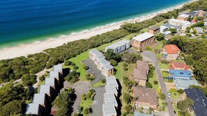 An aerial view of a coastal residential area with a beach, green spaces, and various buildings including a multi-story apartment complex and detached houses. - 'Shoaley Shores on Intrepid'  Overlooking Shoal Bay Beach (Nelson Bay)