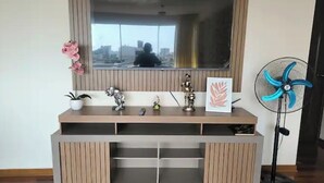 Ein modernes Wohnzimmer mit einem großen an der Wand montierten Fernseher, einer Holz-Unterhaltungseinheit mit Regalen, einer kleinen Topfpflanze und einem Standventilator.