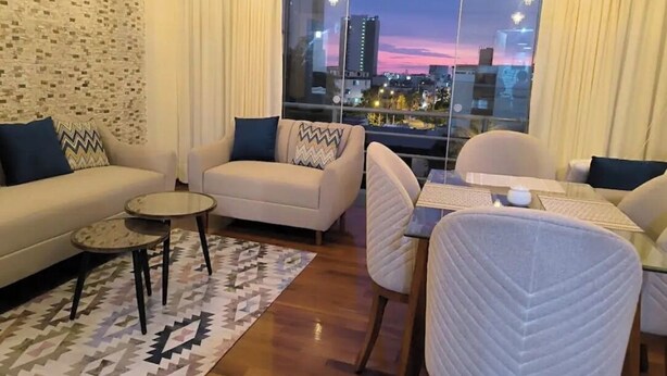 Ein modernes Wohnzimmer mit Sofa, Sesseln, einem kleinen Tisch und Blick auf eine Stadtlandschaft im Abenddämmerung.