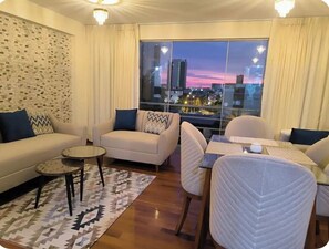 A modern living room with a sofa, armchairs, a small table, and a view of a cityscape at dusk. - 3-bedroom apartment, near Miraflores, San Borja and Surco (Surquillo)