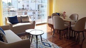 Ein geräumiger Wohnbereich mit einer Couch, einem Couchtisch und einem Esstisch. Große Fenster bieten Blick auf die Stadtlandschaft.