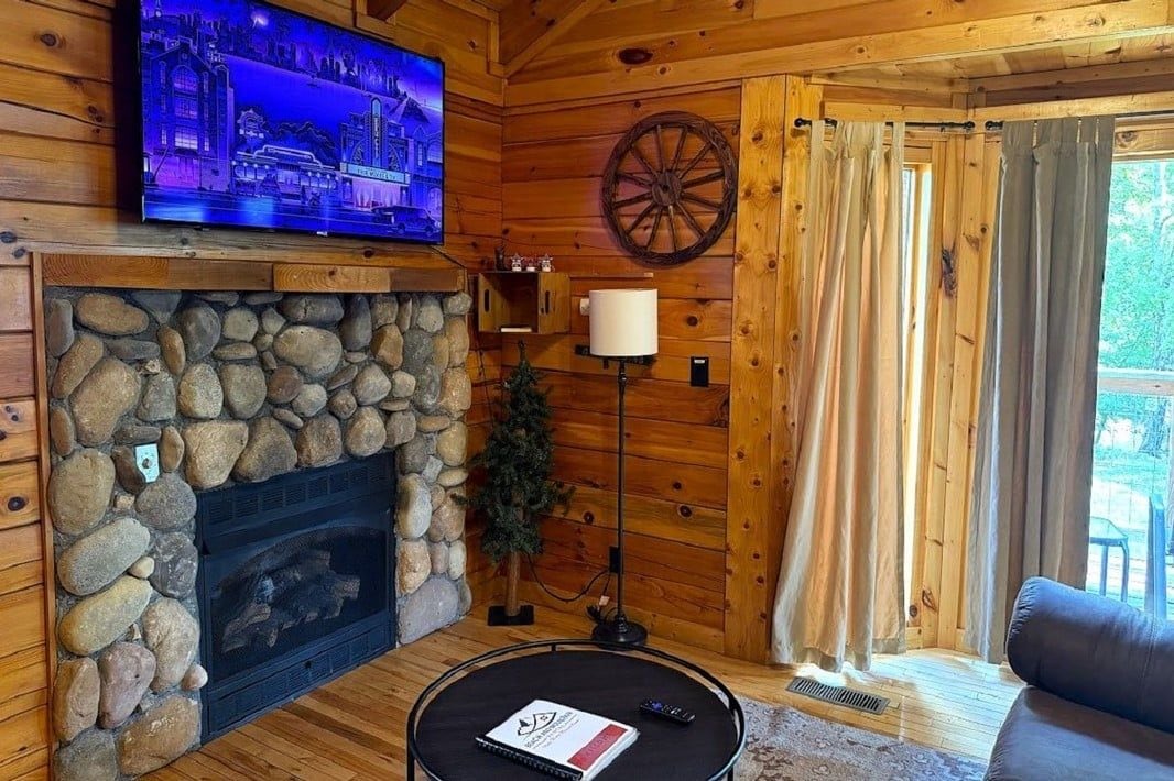 A cozy cabin interior with a stone fireplace, a mounted television, and a large wooden wheel.