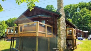 A log cabin with a stone chimney, a wooden deck, and a fenced-in area. - Skinny Dippin: Free Tickets, Pet Friendly, Creek (Pigeon Forge)