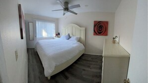 Ein Schlafzimmer mit einem Bett, einem Deckenventilator, einem Nachttisch und einem Fenster mit Jalousien. An der Wand hängt ein abstraktes, rotes Gemälde.