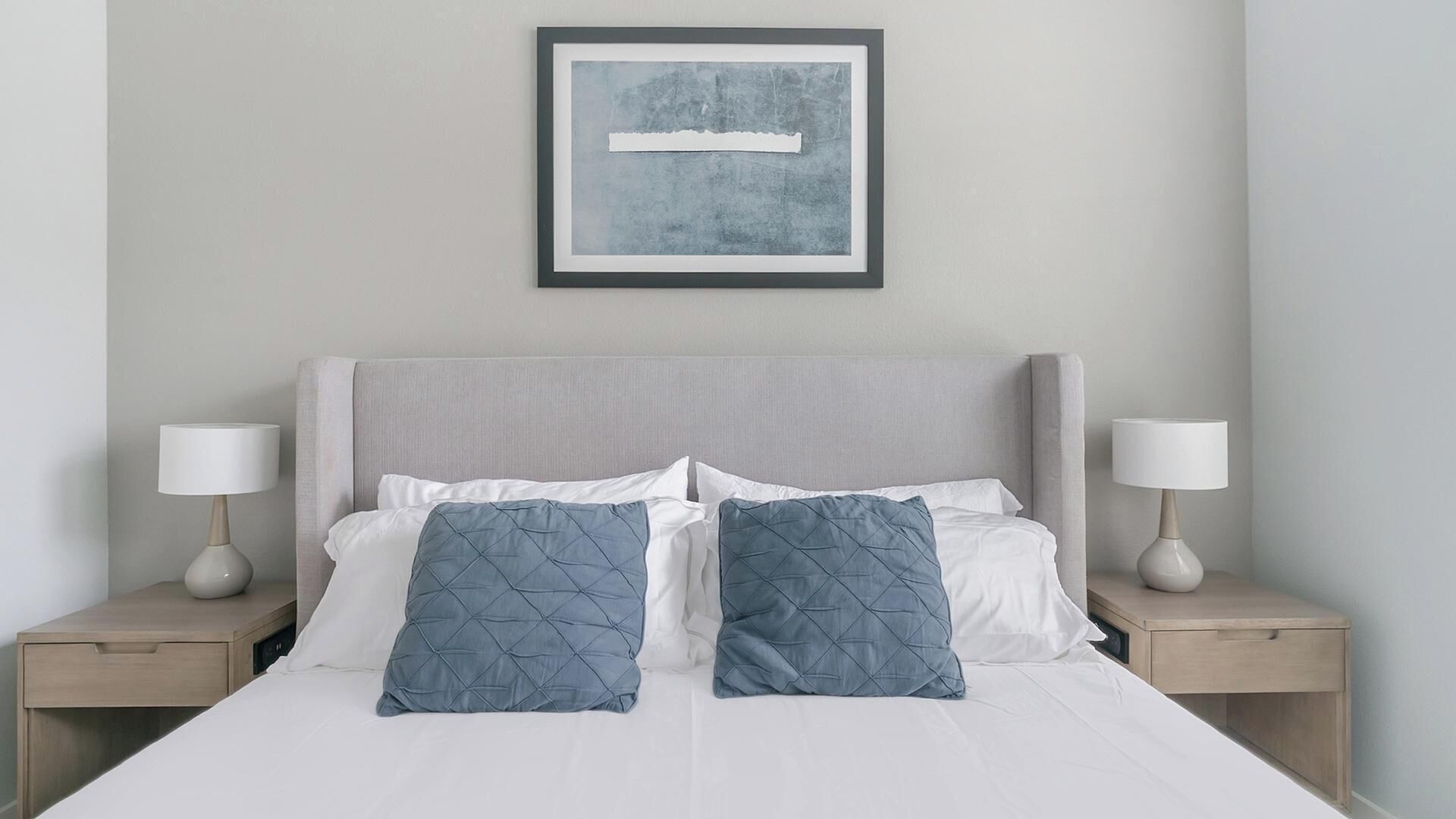 Een net opgemaakt bed met witte lakens en twee blauwe kussens, zijkanten omlijst door twee nachtkastjes met lampen, in een kamer met een effen muur en een kunstwerk boven het bed.