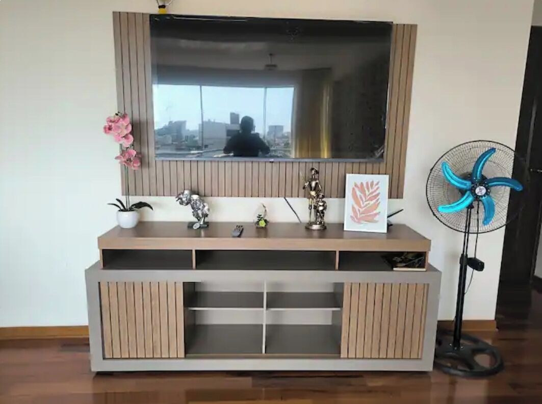 Un salon moderne avec un grand téléviseur fixé au mur, un meuble de rangement en bois doté d’étagères, une petite plante en pot et un ventilateur sur pied.