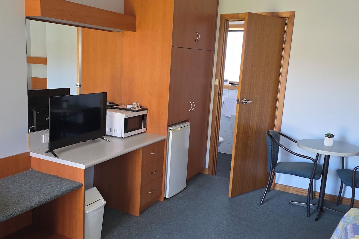 A compact room with a desk, a television, a small refrigerator, a trash bin, and a dining area with two chairs and a small table.