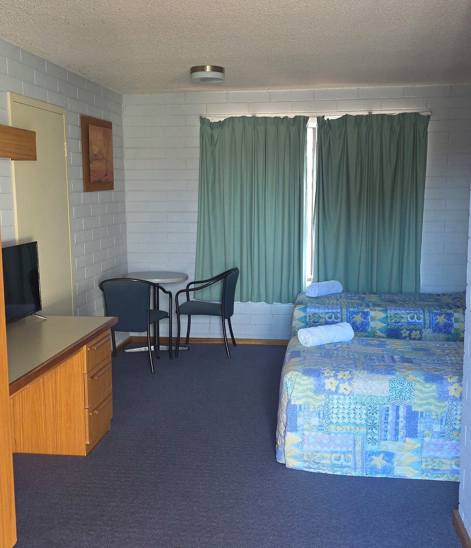 A room with a wooden desk, two chairs, a bed with a patterned duvet, and a window with green curtains.