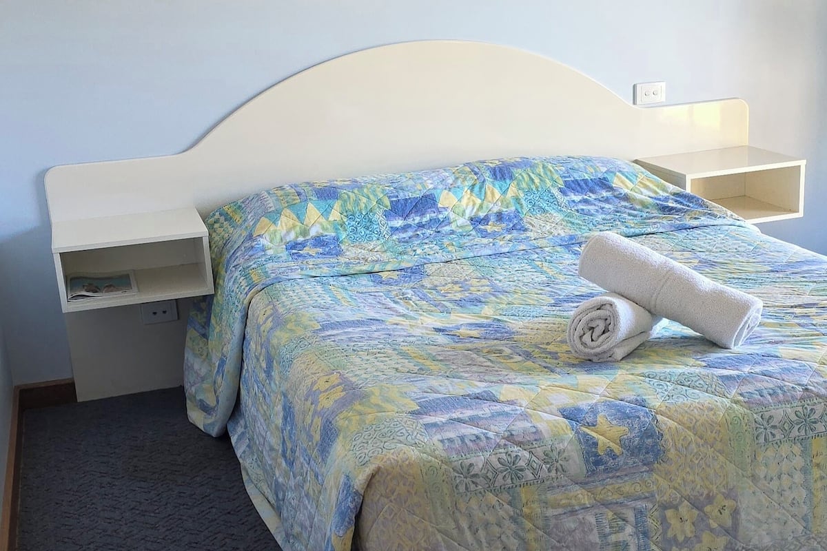 A neatly made bed with a patterned bedspread, a rolled towel, and a white headboard.
