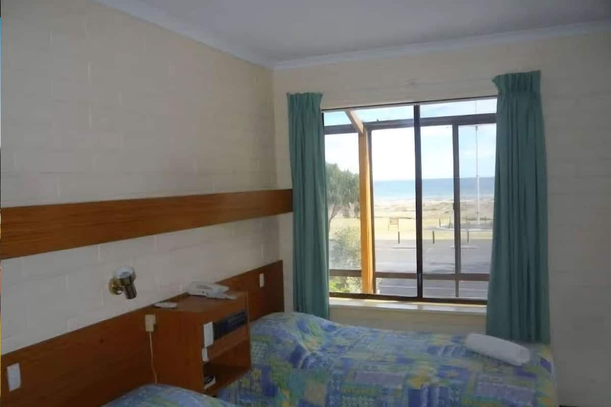 A room with two beds, a wooden headboard, a window with curtains, and a view of the beach.