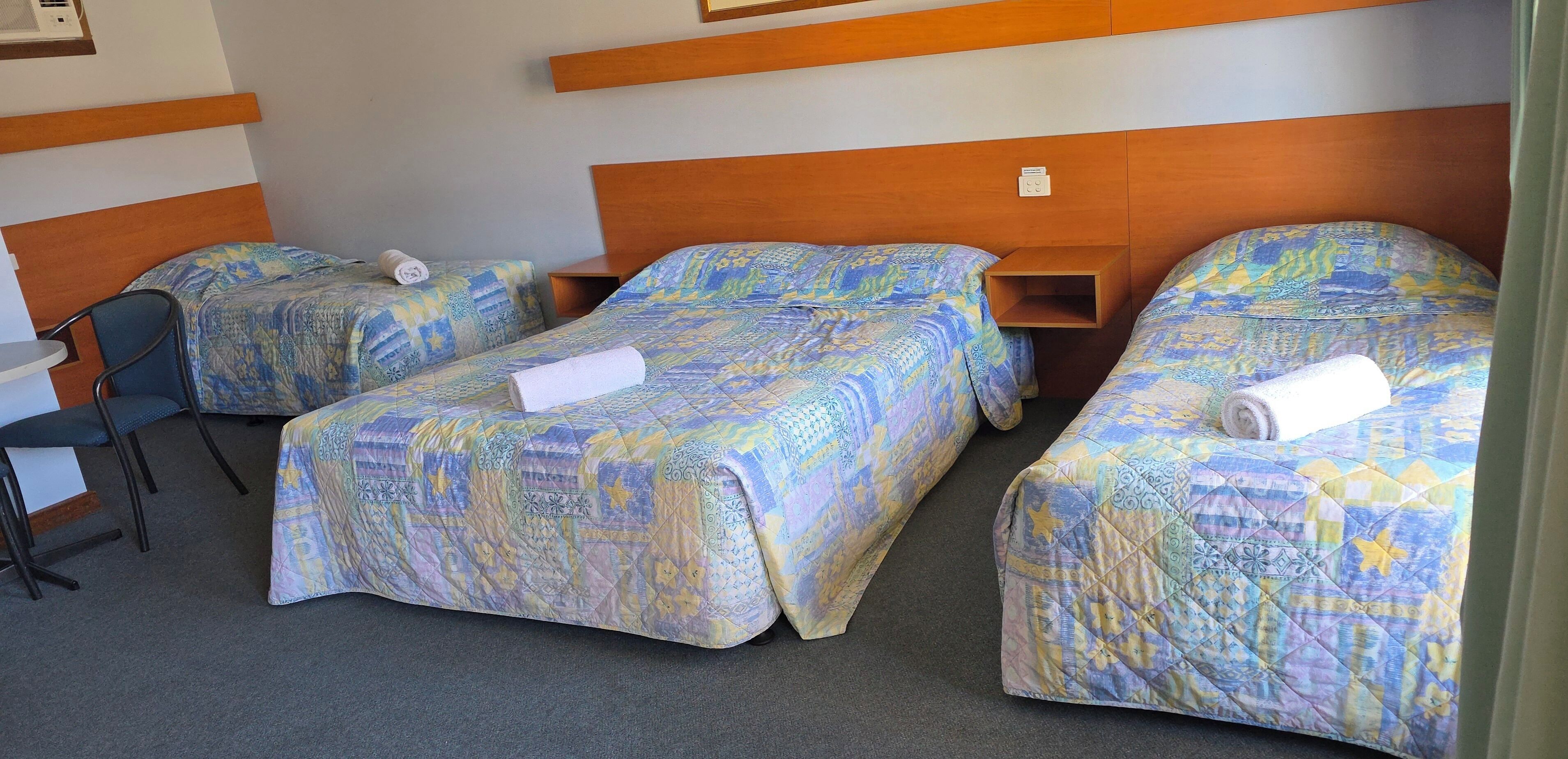 Habitación de hotel con tres camas, cada una con colcha estampada. Hay una silla y una mesita de noche con lámpara.