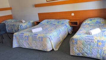 Habitación de hotel con tres camas, cada una con colcha estampada. Hay una silla y una mesita de noche con lámpara.