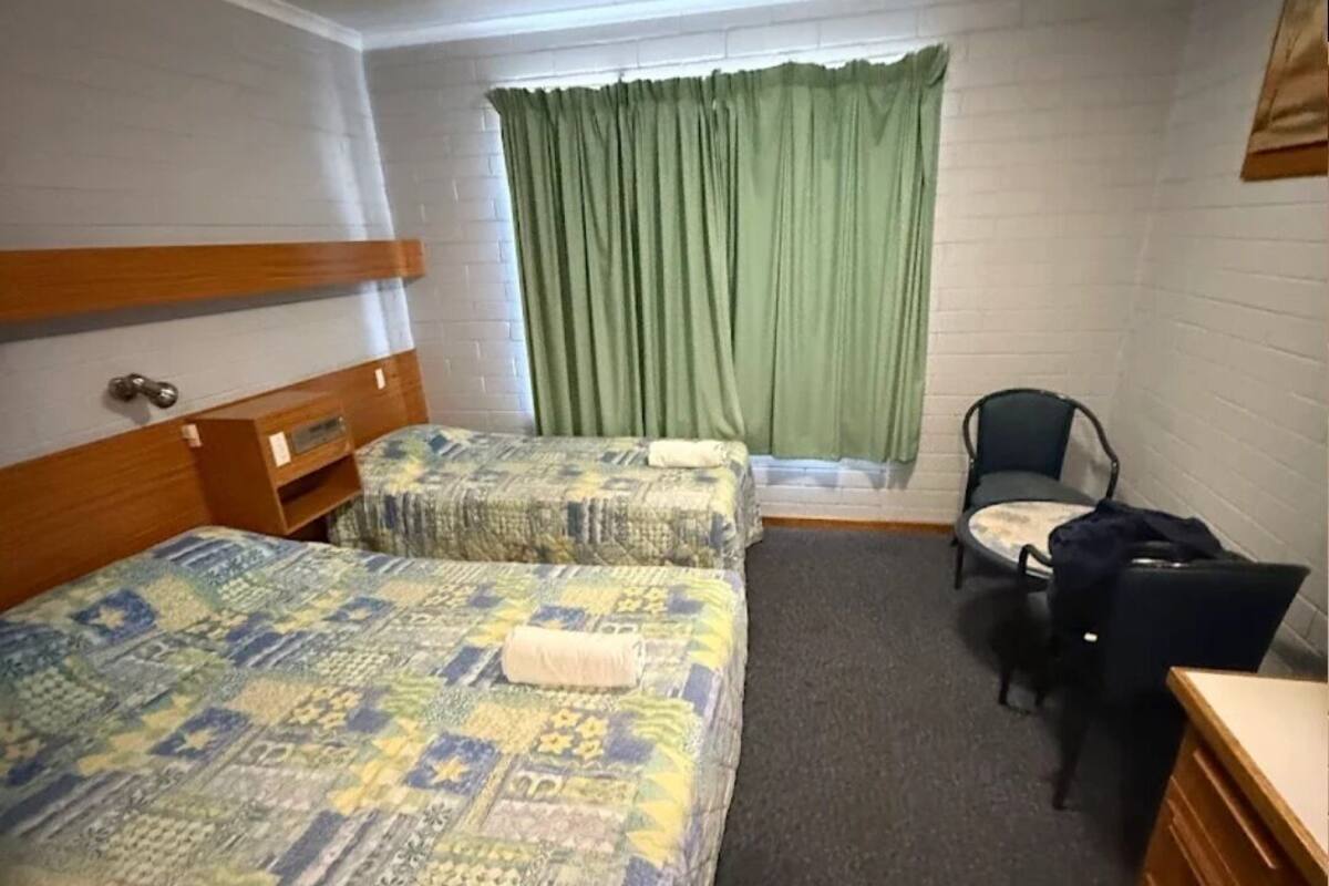 A hotel room with two beds, a desk, a chair, and a window with green curtains.